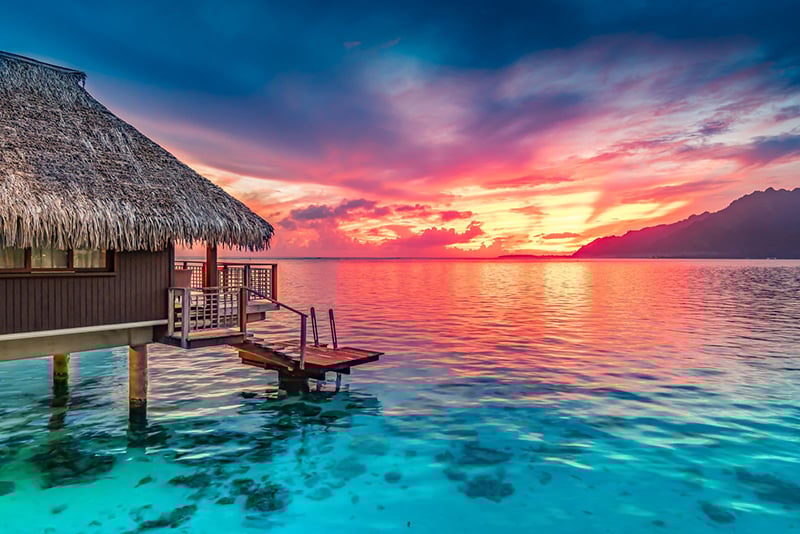 Colorful sunset in the background of a vacation hut above turquoise water.